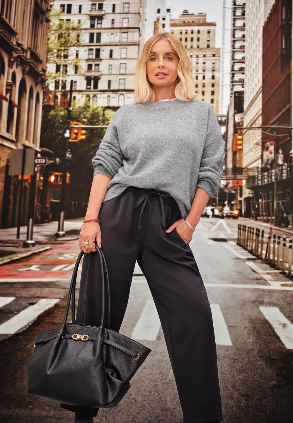 Model in a grey jumper and black trousers from Peacocks, carrying a black bag on a city street